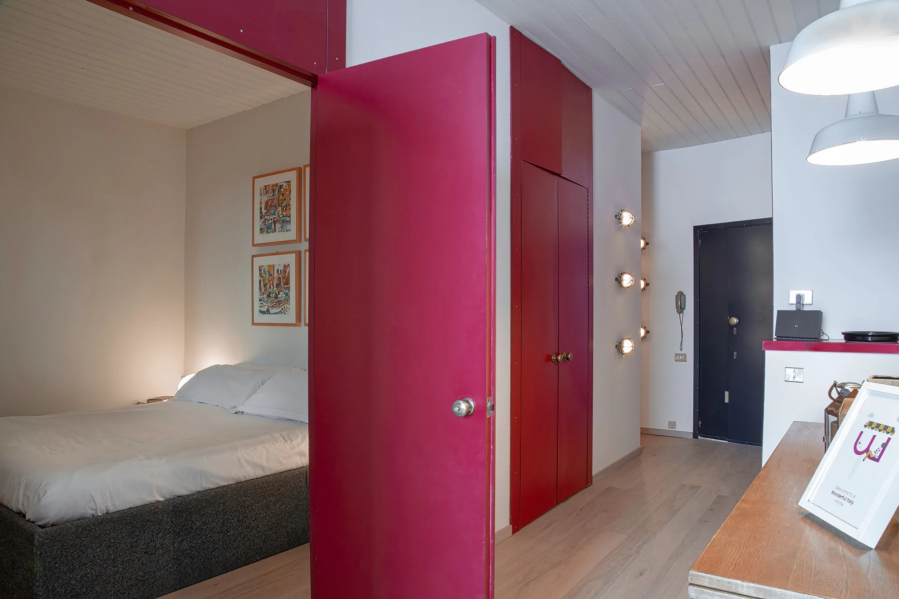 Se ve un dormitorio con una cama junto a una puerta magenta y un pasillo.