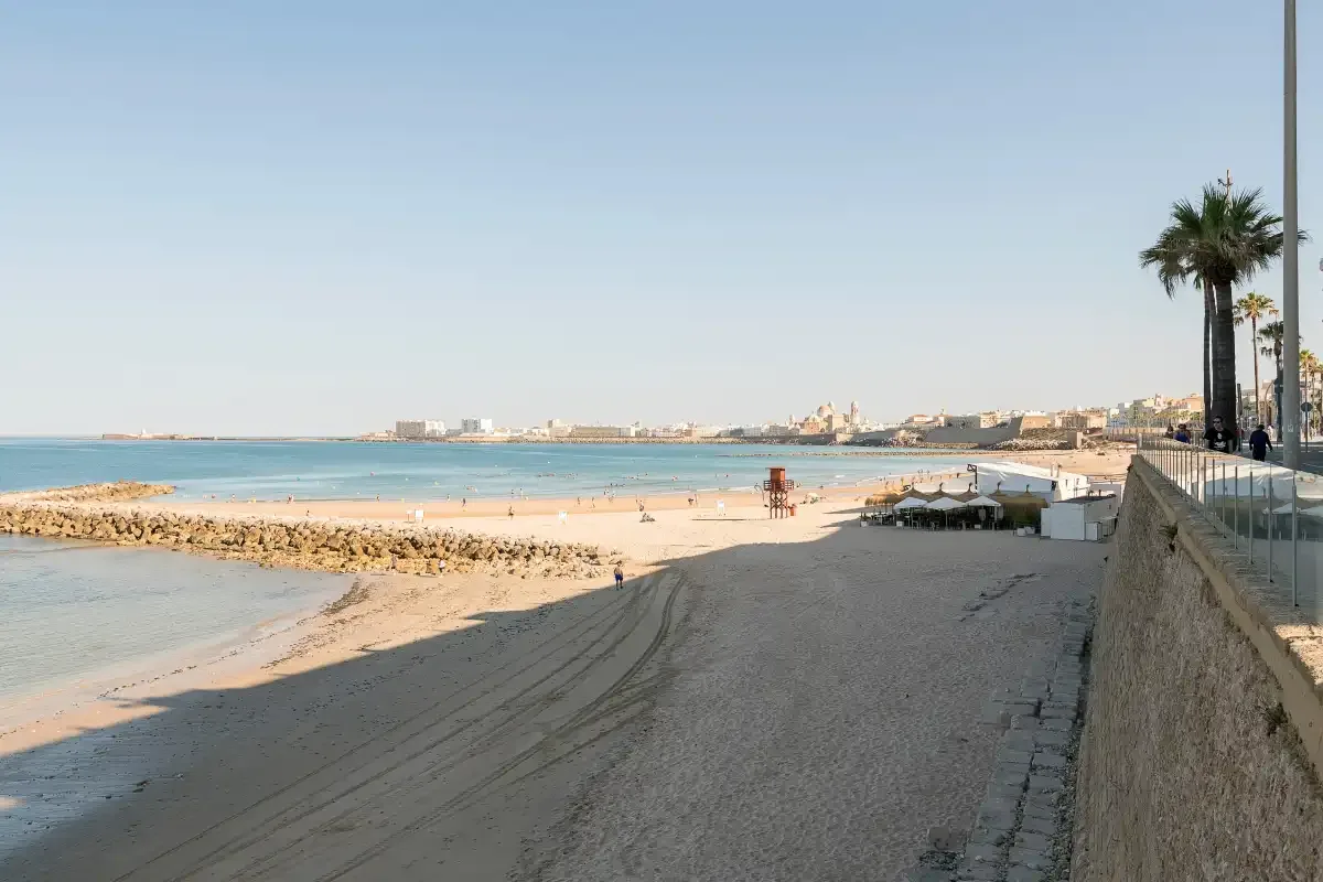 A sandy beach curves beside calm blue water, with a promenade along the shore.