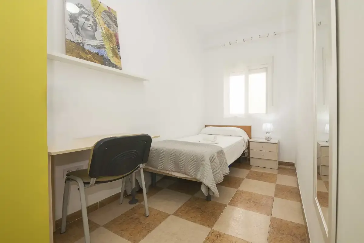 A bedroom with a bed, desk, and chair beside a bright window.