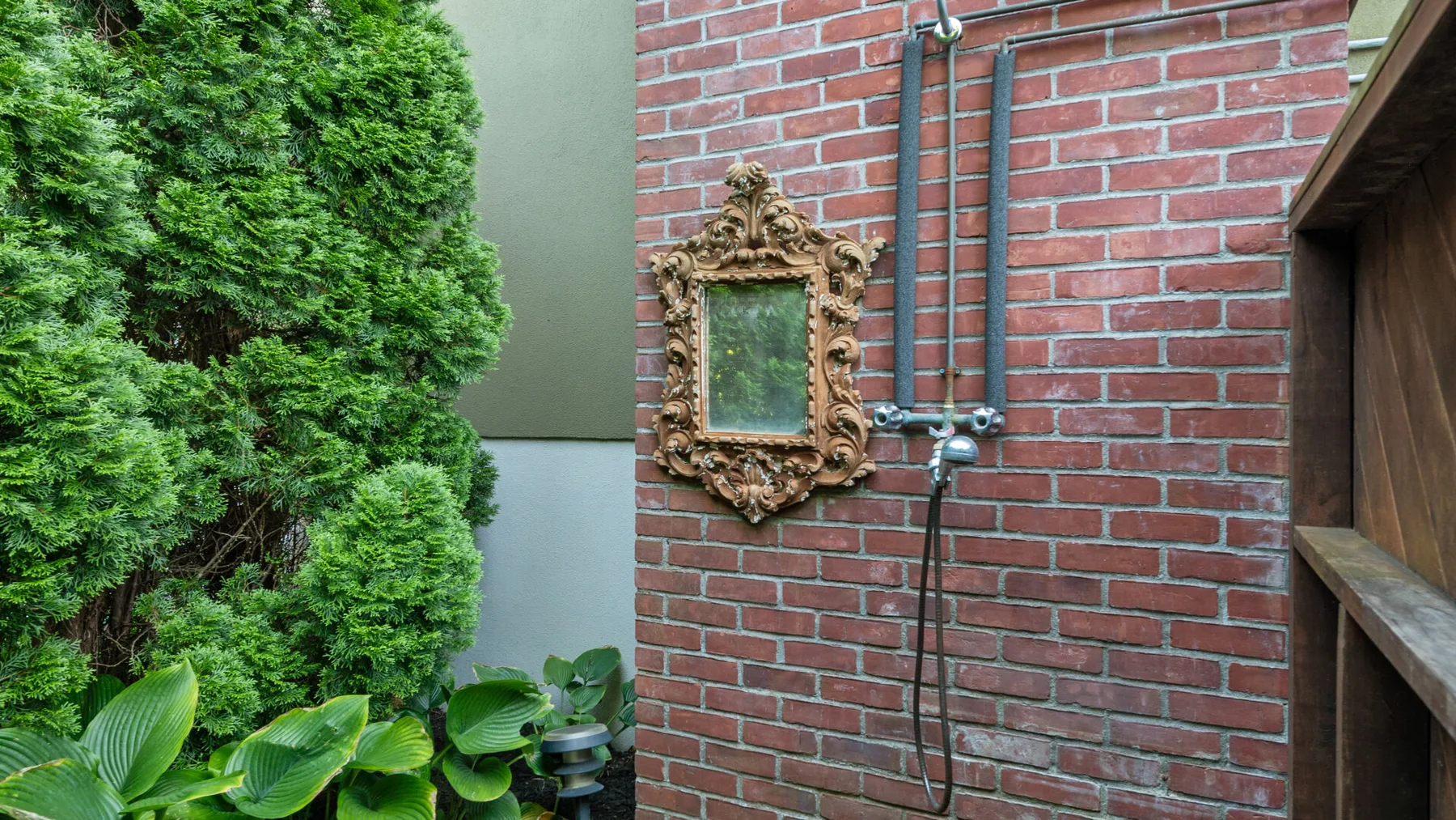 Una pared exterior de ladrillo con una ducha y un espejo ornamentado junto a vegetación.