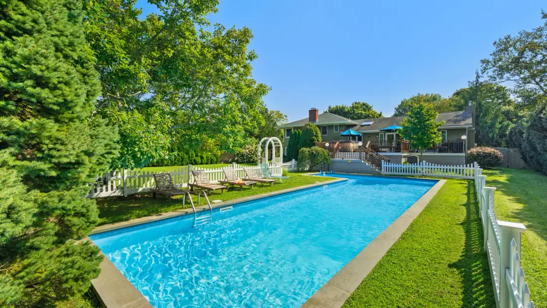 An outdoor swimming pool sits beside a fenced lawn and leafy trees.