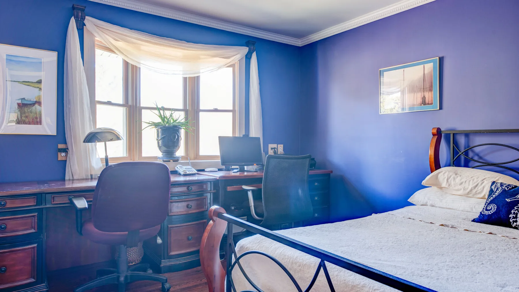 Un dormitorio azul tiene una cama y un escritorio junto a una ventana luminosa.