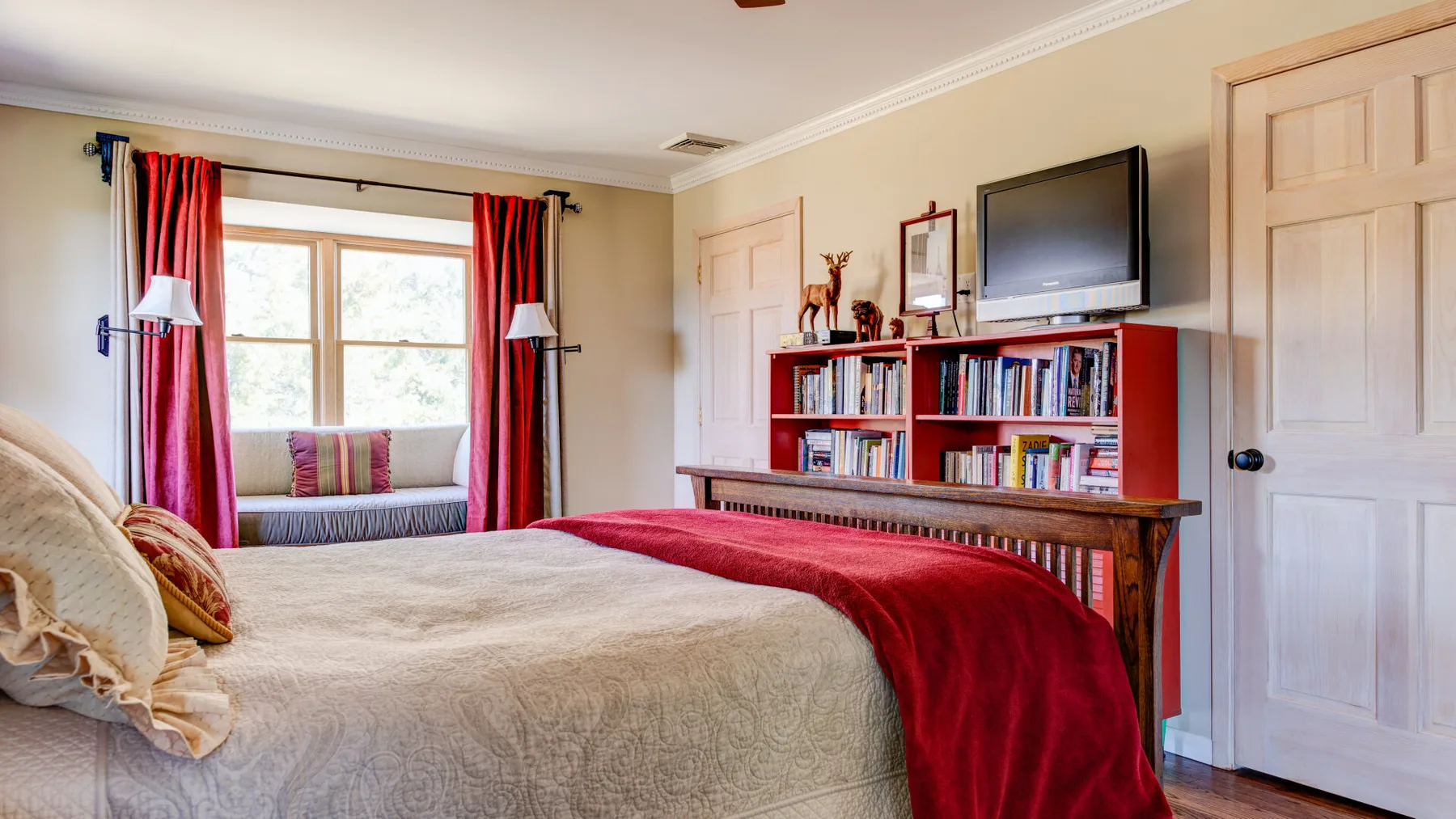 Un dormitorio con una cama, cortinas rojas y una estantería bajo un televisor.