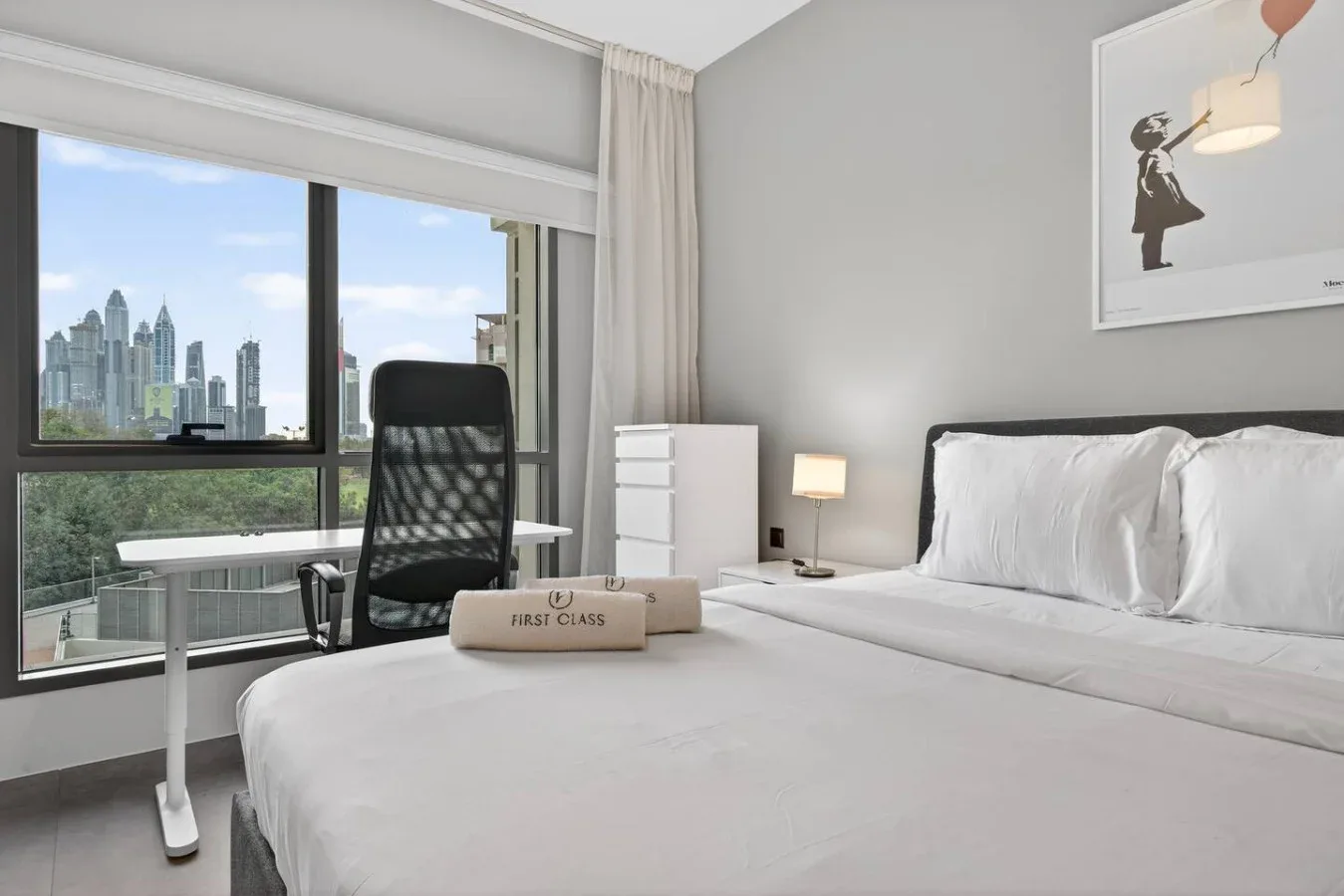 Un dormitorio luminoso con una cama, un escritorio y grandes ventanas con vistas a edificios.