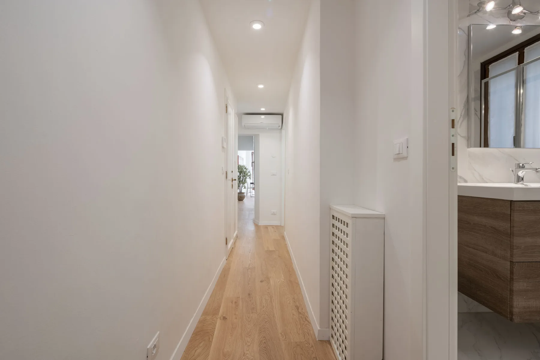 A narrow hallway with light wood flooring leads to a bright room.
