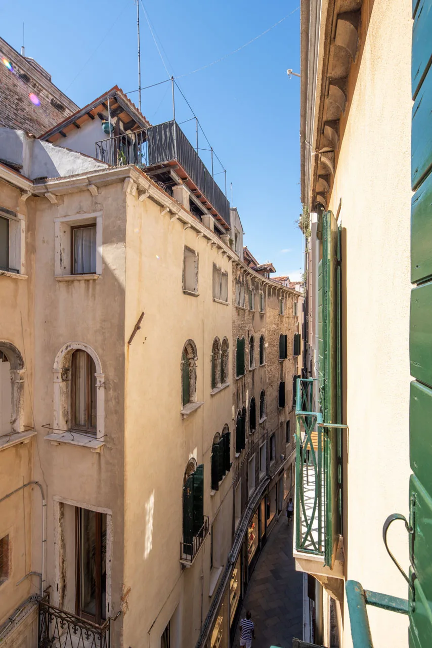 A narrow Venice street runs between pale buildings, viewed from a green-shuttered window.