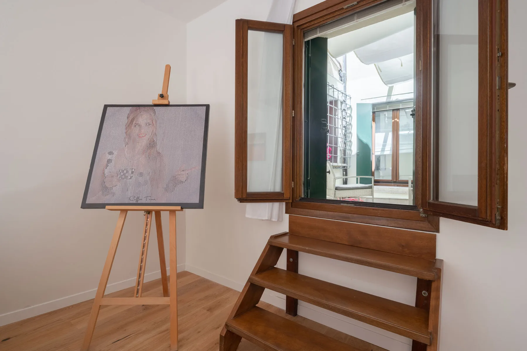 A room with an open wooden-framed window, steps, and a portrait on an easel.