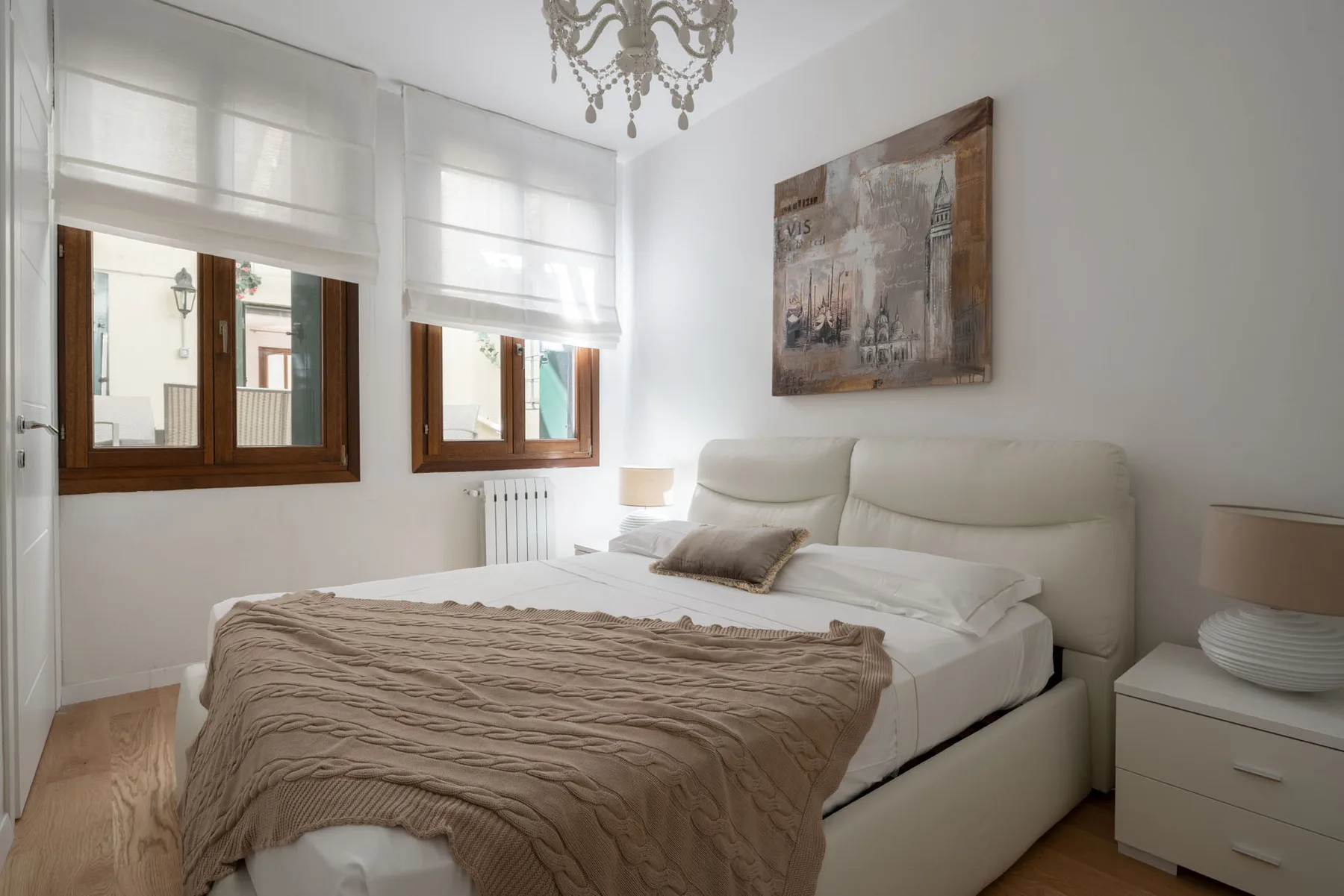 A bright bedroom with a bed, two windows, and a framed wall print.