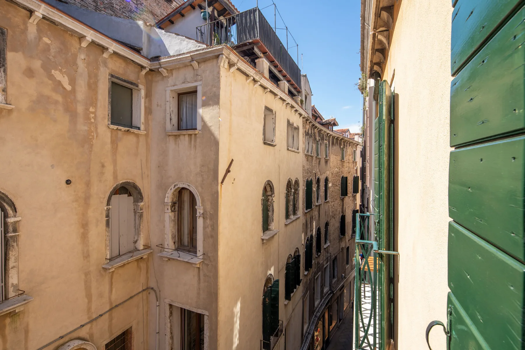 A narrow street runs between sunlit buildings with shuttered windows.