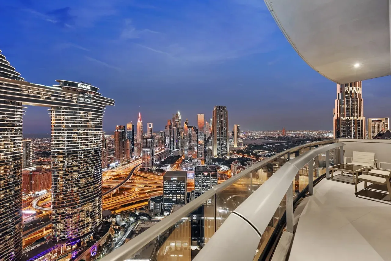 A balcony overlooks a city skyline at dusk, with seating along the railing.