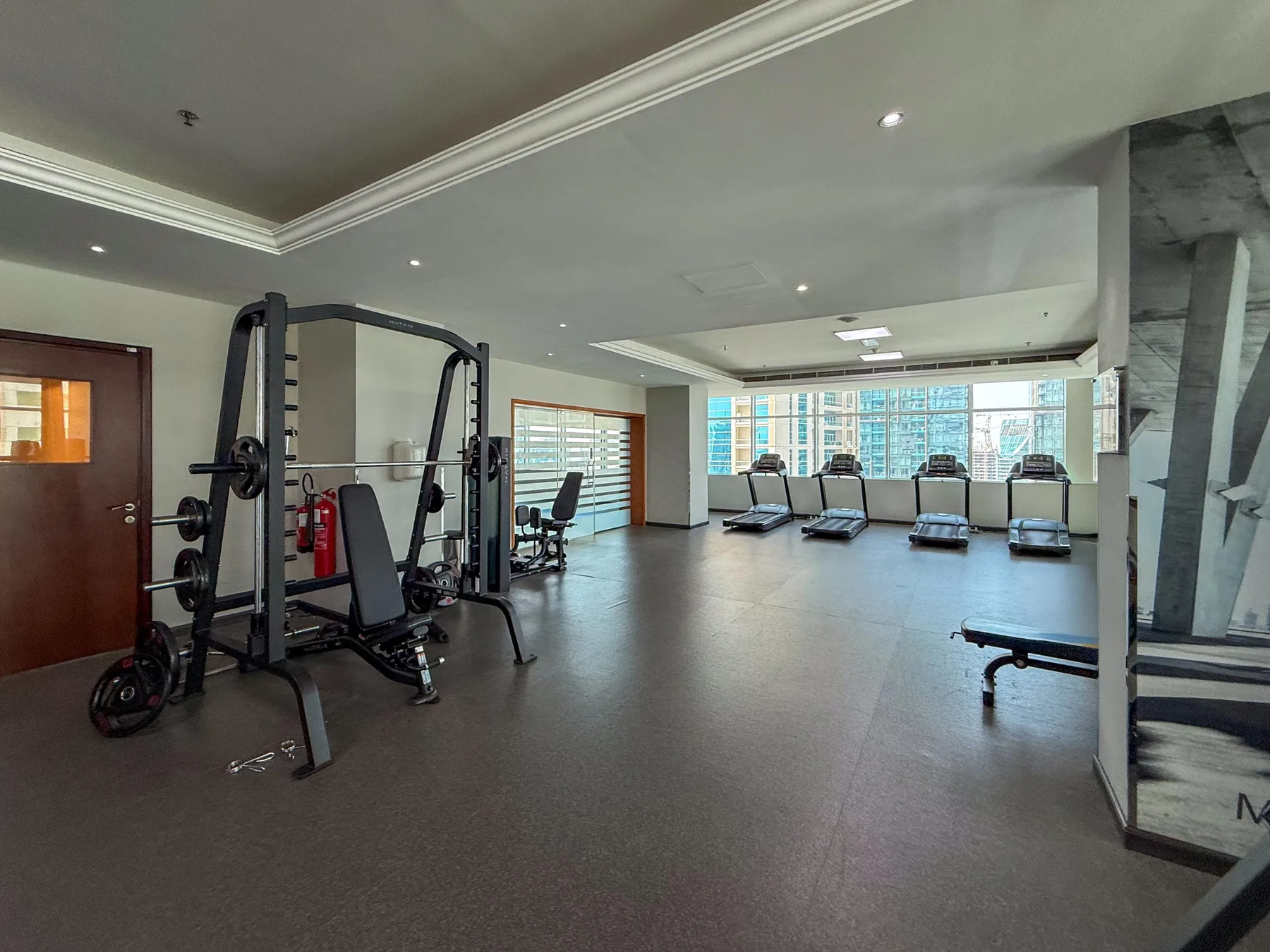 Un gimnasio con máquinas de ejercicio y cintas de correr frente a grandes ventanas.