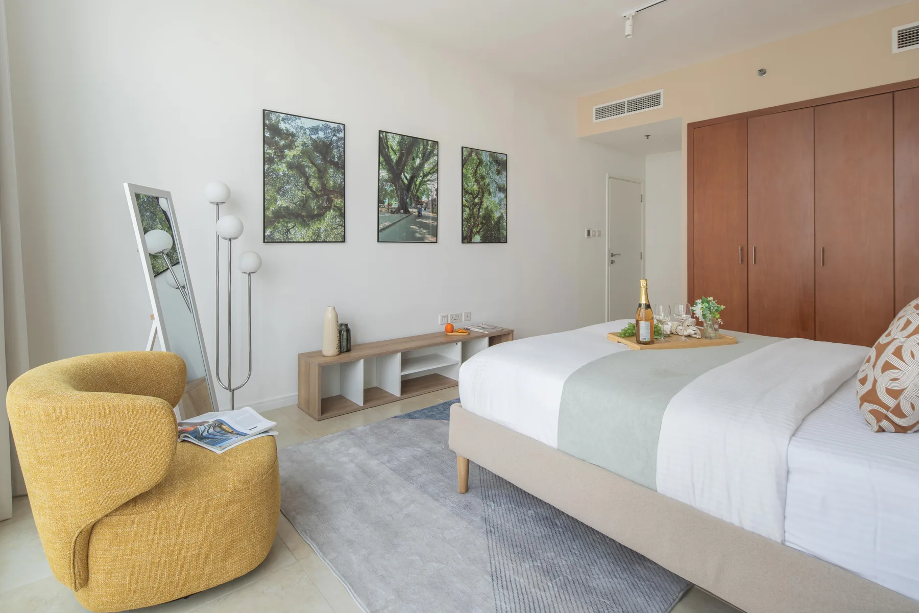 Un dormitorio luminoso con una cama tendida, un sillón amarillo y cuadros enmarcados.