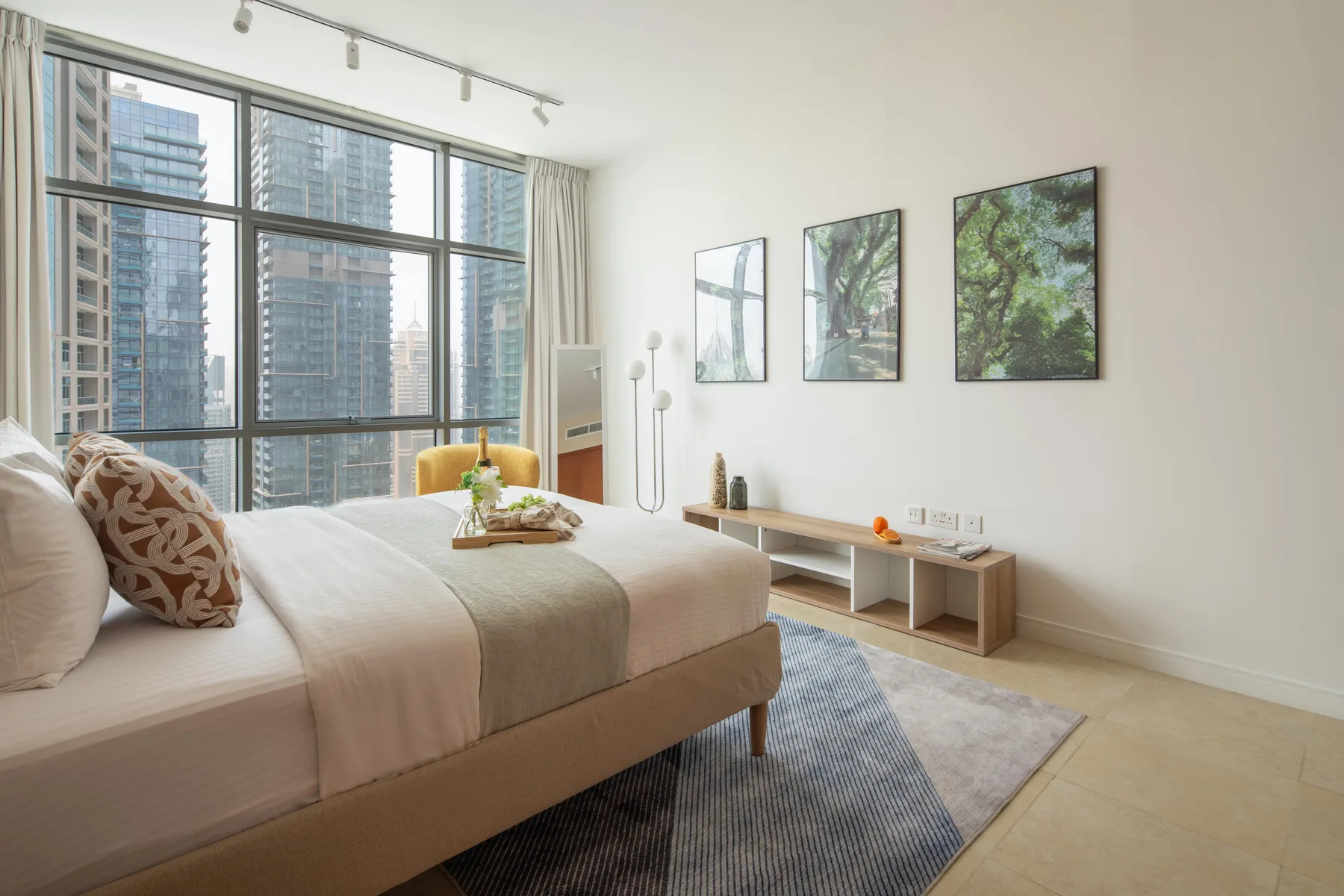 Un dormitorio luminoso con una cama junto a grandes ventanas con vistas a edificios cercanos.