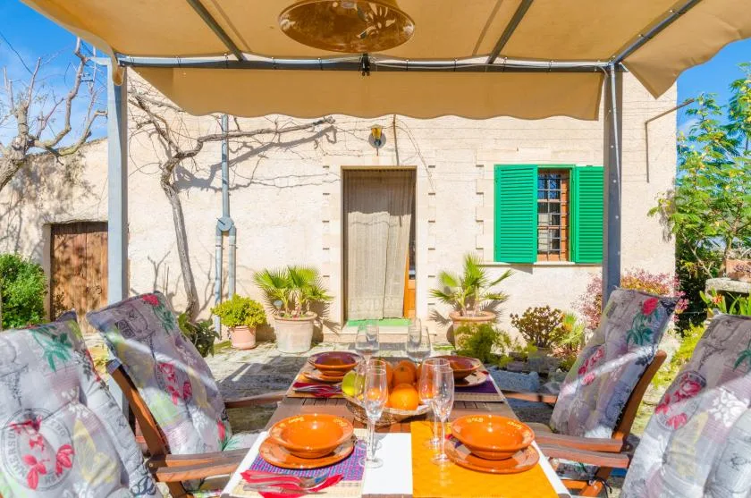 A shaded outdoor dining area sits in front of a pale house with green shutters.