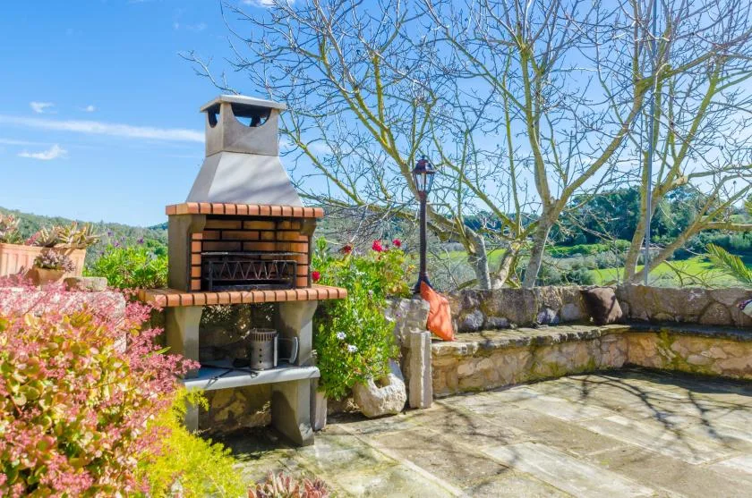An outdoor patio features a brick barbecue beside a stone wall and flowering plants.