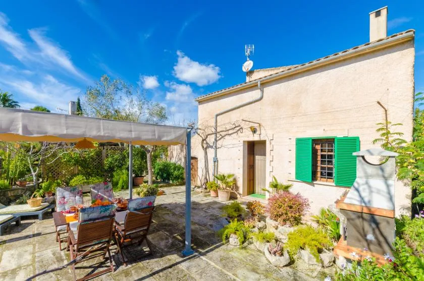 A sunny patio beside a pale house, with shaded seating and garden plants.