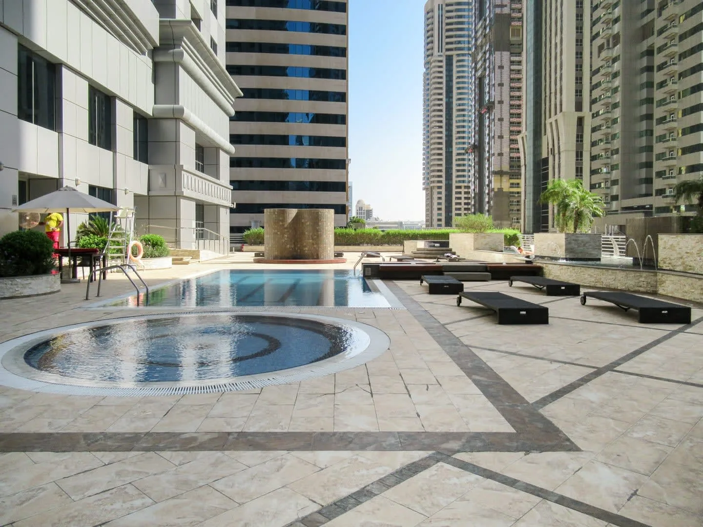Una piscina al aire libre y una fuente circular están entre edificios altos.