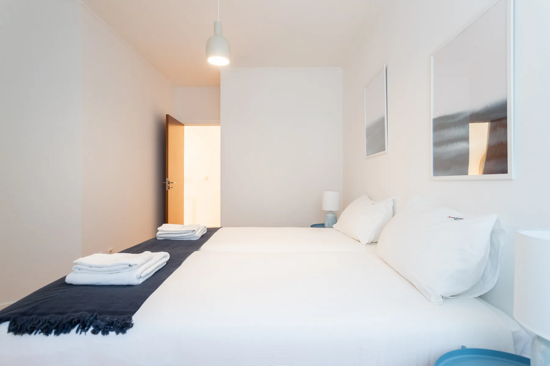 Un dormitorio luminoso con una cama blanca, una manta oscura y la puerta abierta.
