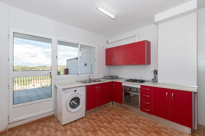 Una cocina luminosa con armarios rojos, una lavadora y una puerta al balcón.