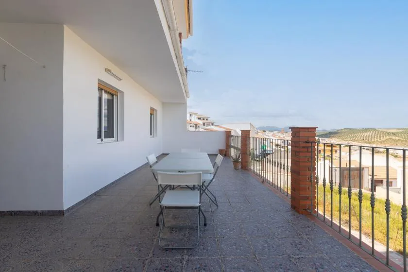 Una terraza exterior embaldosada tiene una mesa y asientos junto a una barandilla metálica.