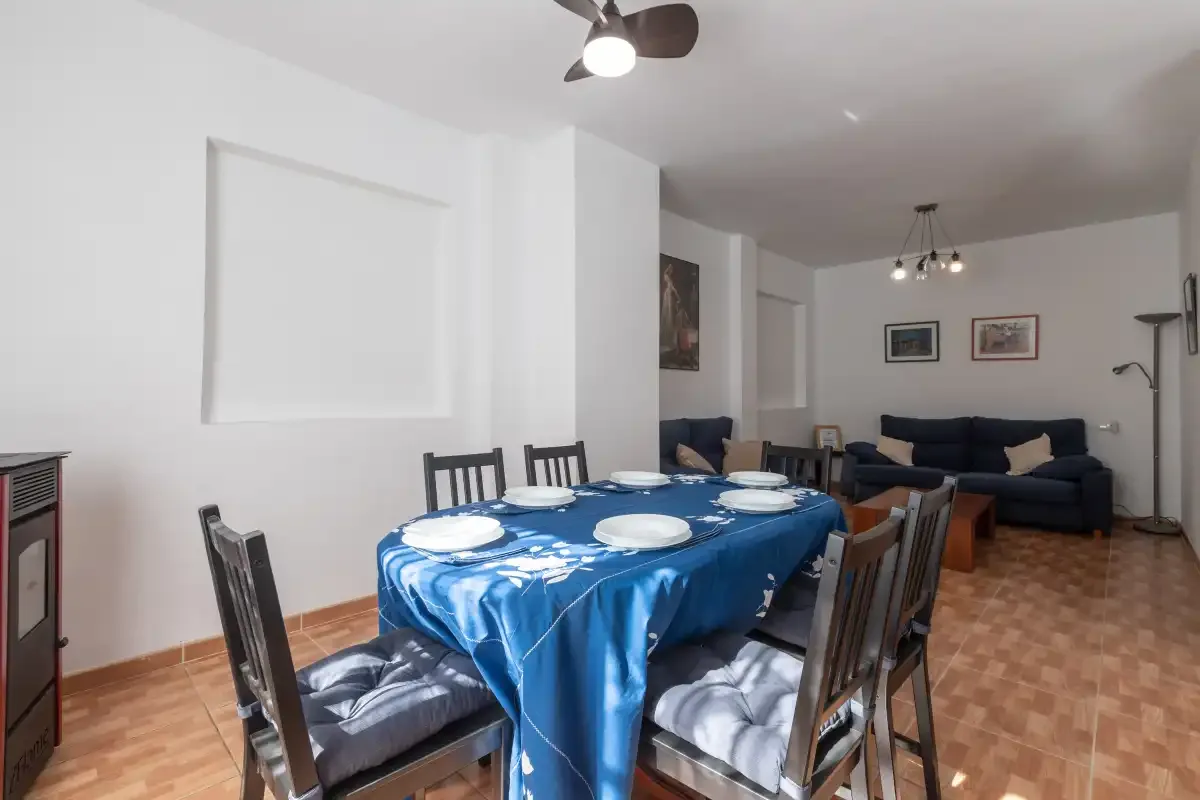 Comedor con una mesa cubierta de azul y asientos, abierto a una zona de estar.