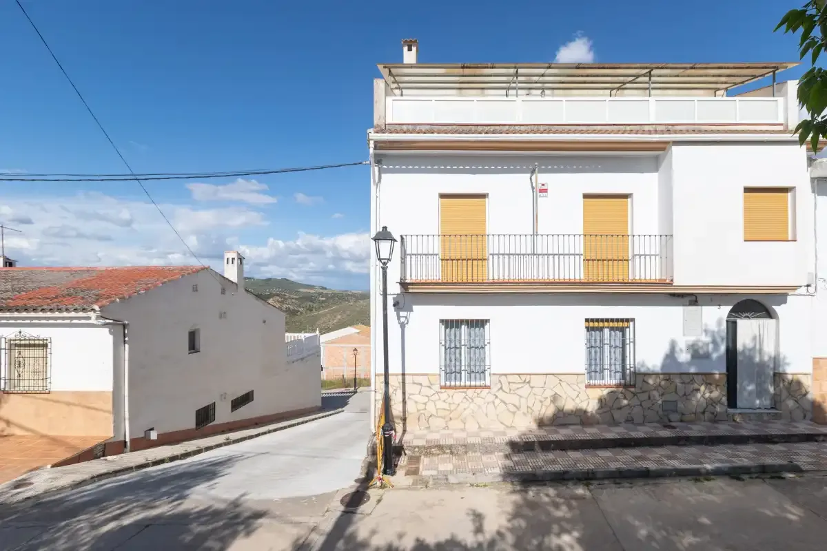 Un edificio blanco de varias plantas se encuentra junto a una calle estrecha en un pueblo soleado.