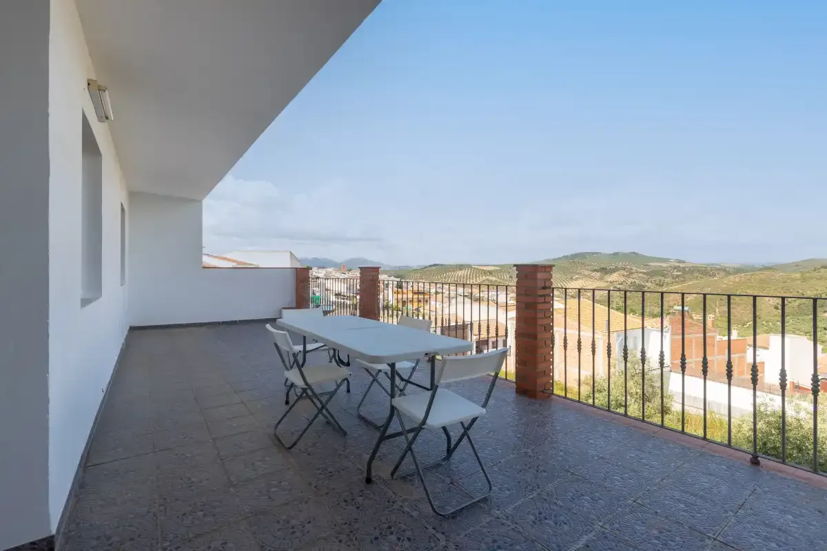 Una terraza exterior cubierta tiene una mesa y asientos junto a una barandilla metálica.