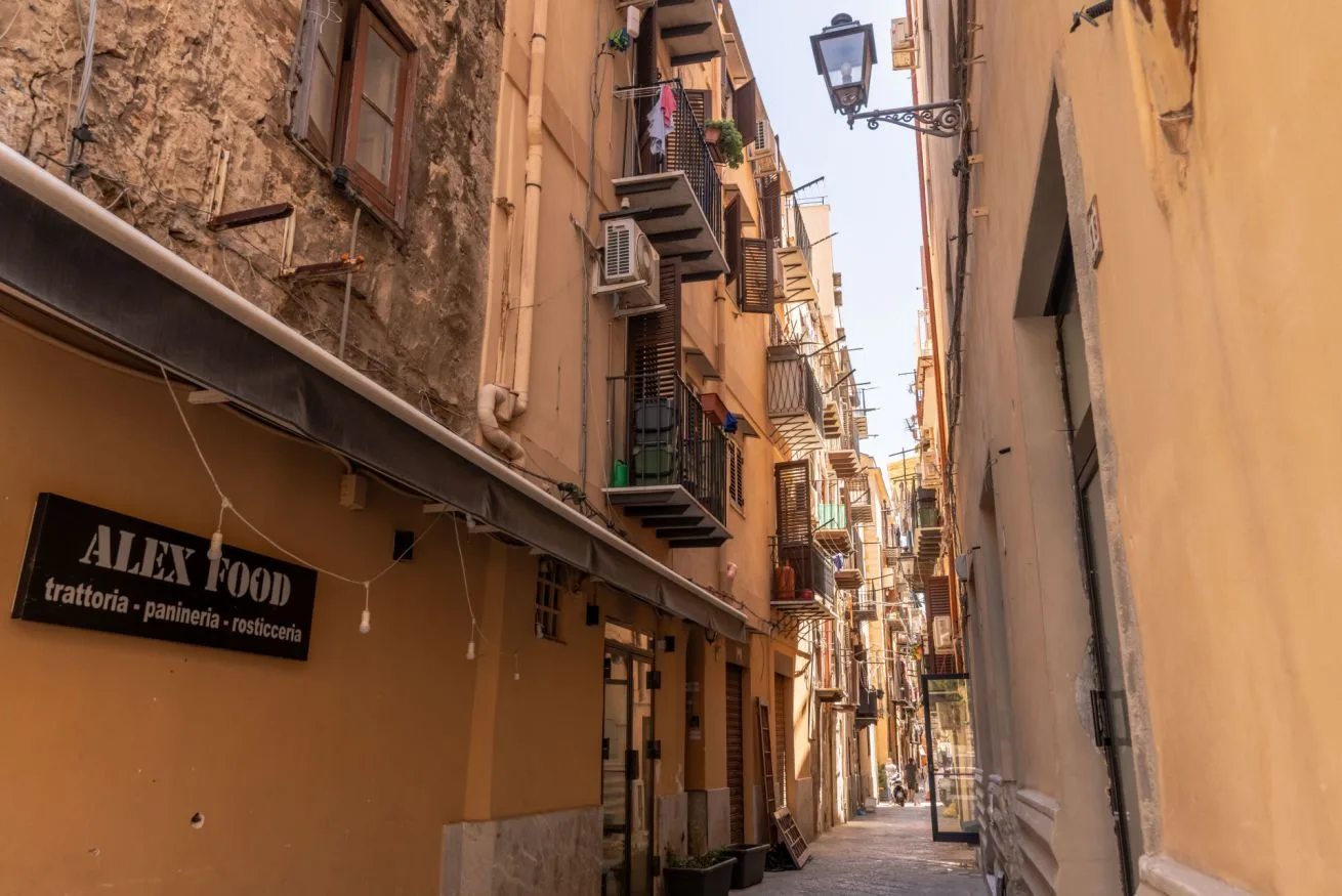 A narrow pedestrian alley runs between closely spaced buildings, with a shop sign and balconies.