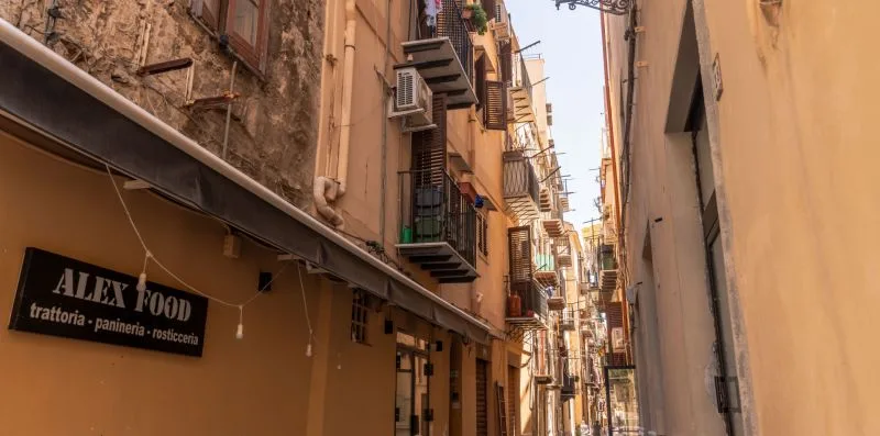 Una calle estrecha pasa entre edificios muy próximos, con un letrero de restaurante a la izquierda.