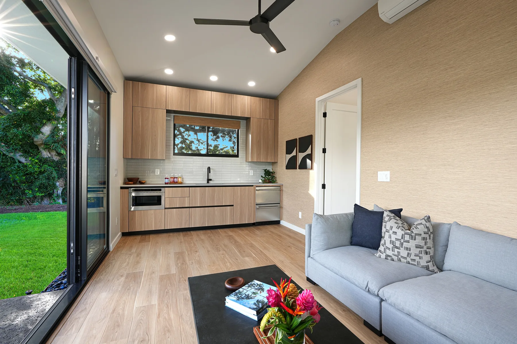 A bright living area with a compact kitchen, a sofa, and sliding glass doors opening onto greenery.