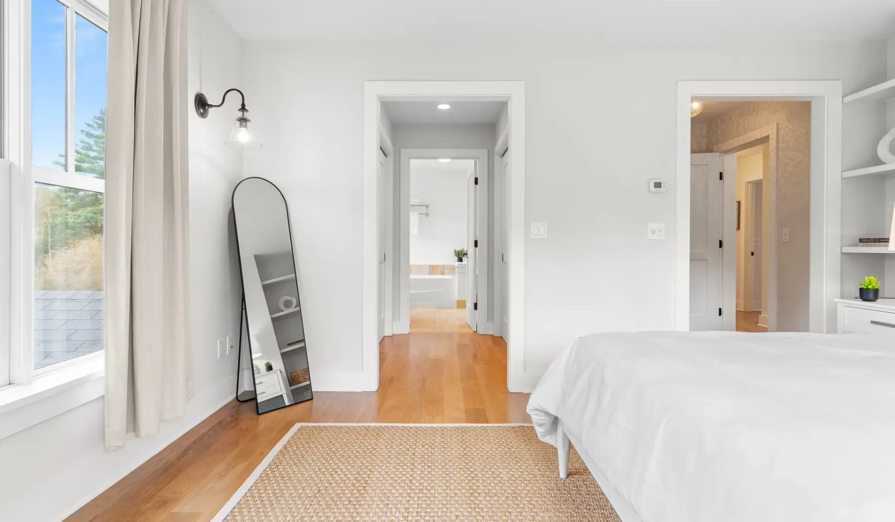 Un dormitorio luminoso con una cama, un espejo de cuerpo entero y una ventana.