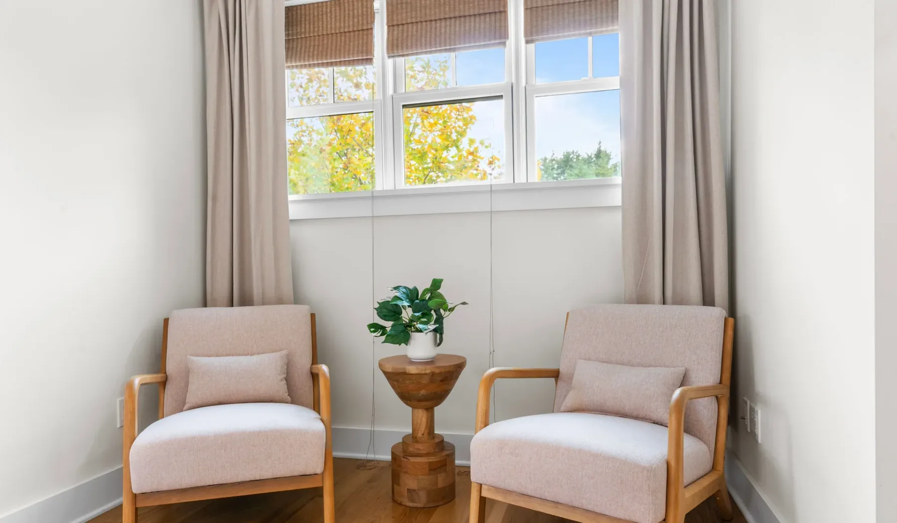 Una luminosa zona de estar tiene dos asientos tapizados junto a una mesa pequeña y ventanas amplias.