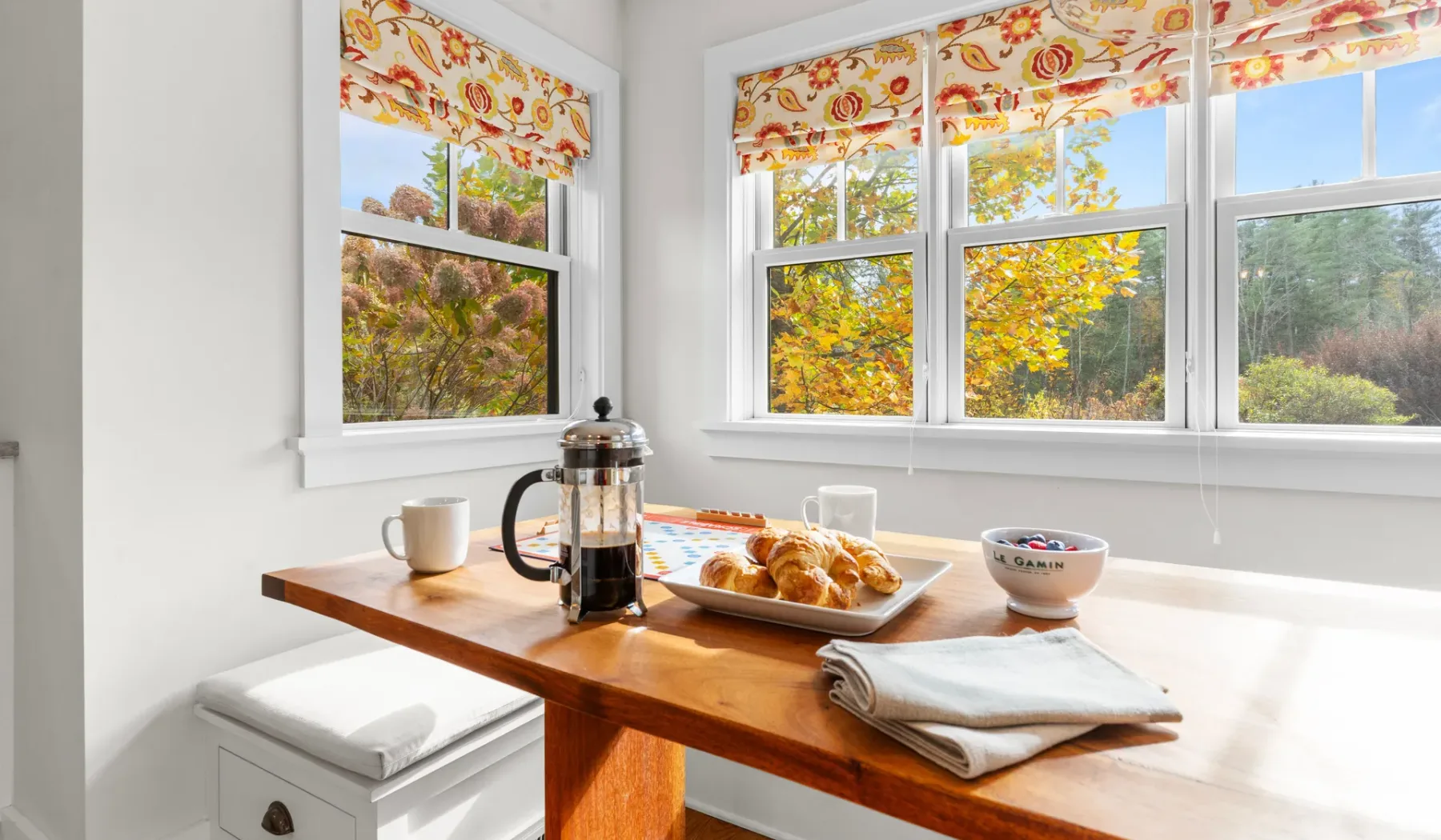 Una mesa de desayuno está junto a ventanas luminosas con vistas a árboles otoñales.