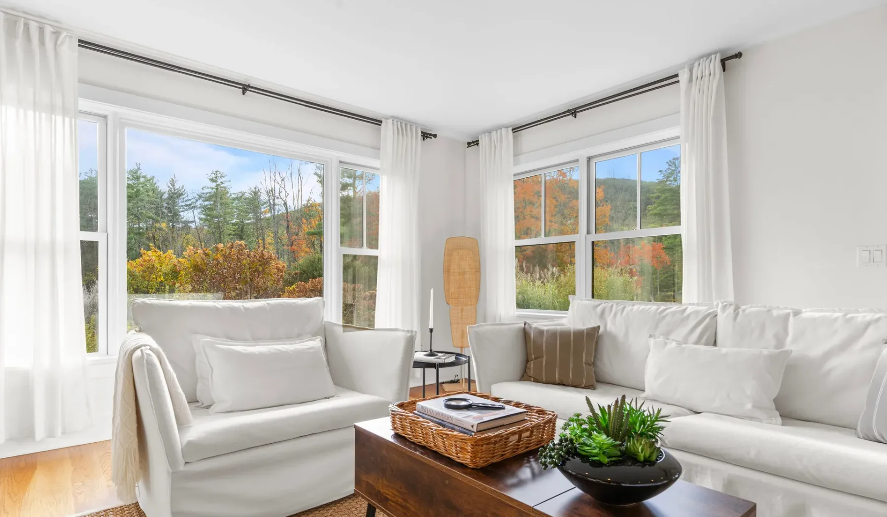Una sala luminosa con asientos blancos y grandes ventanas en esquina con vistas a los árboles.