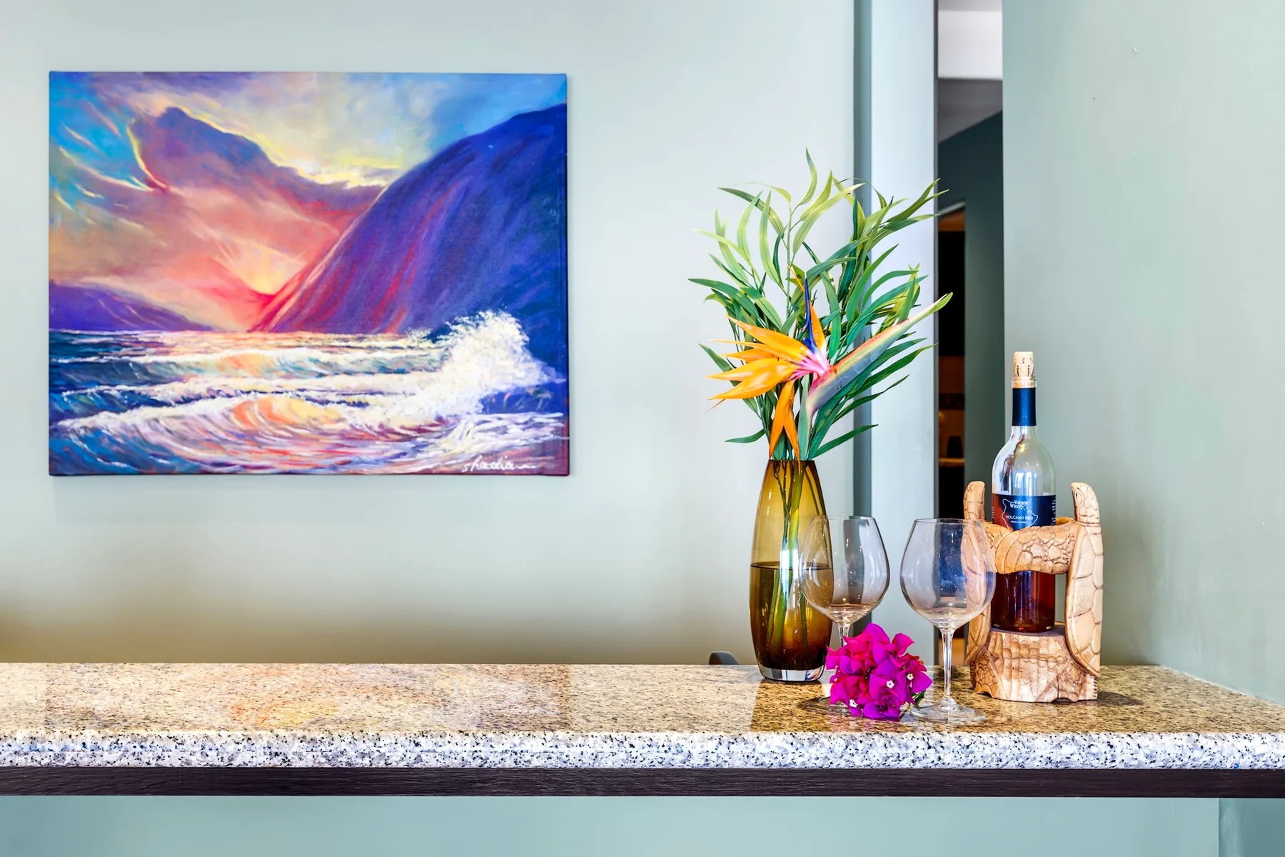 Una colorida pintura marina cuelga sobre una encimera con flores y una botella.