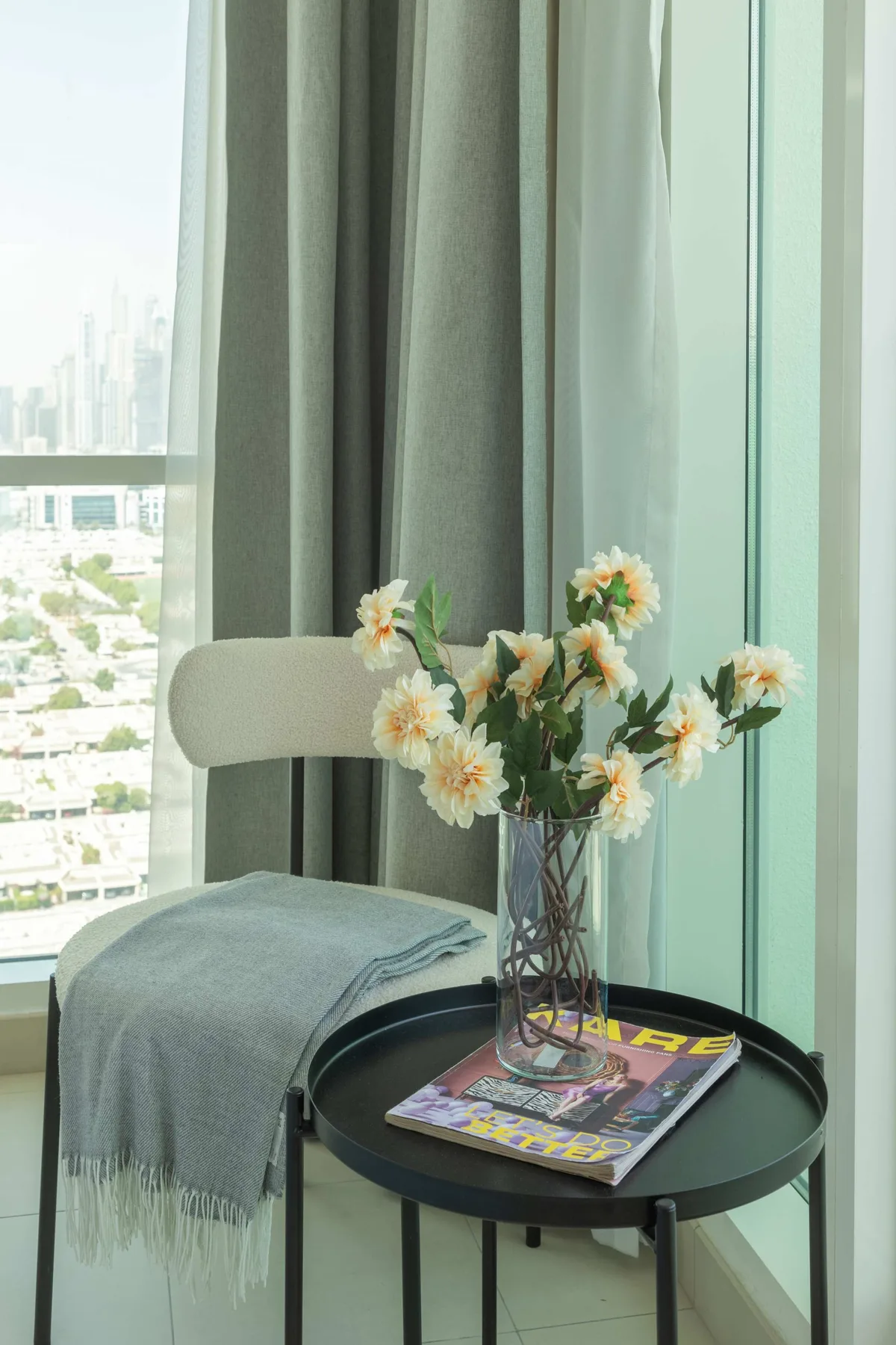 Una mesa pequeña y redonda junto a una ventana tiene un jarrón con flores y revistas.