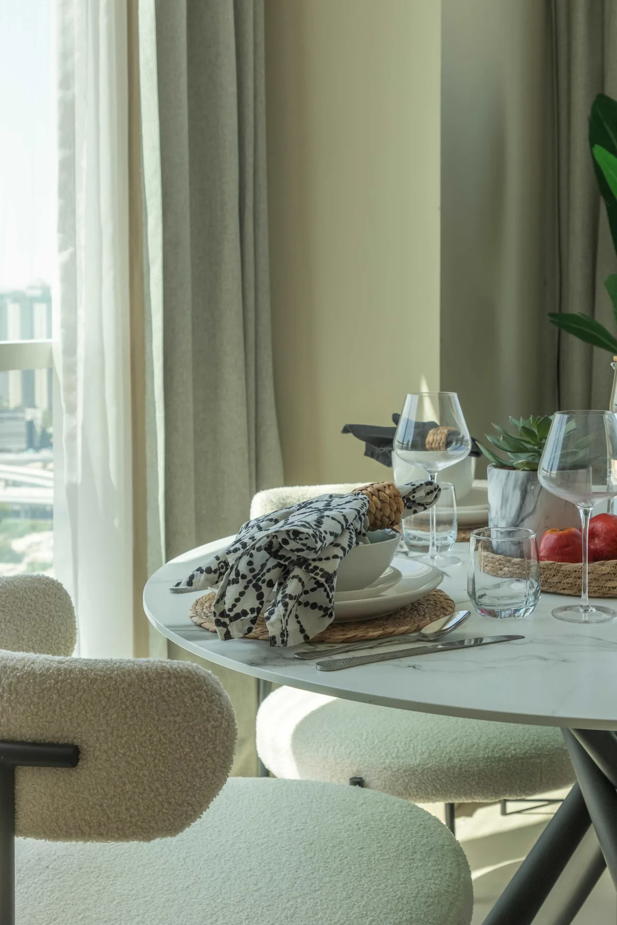 Una mesa de comedor está puesta junto a una ventana con cortinas translúcidas y una planta frondosa.