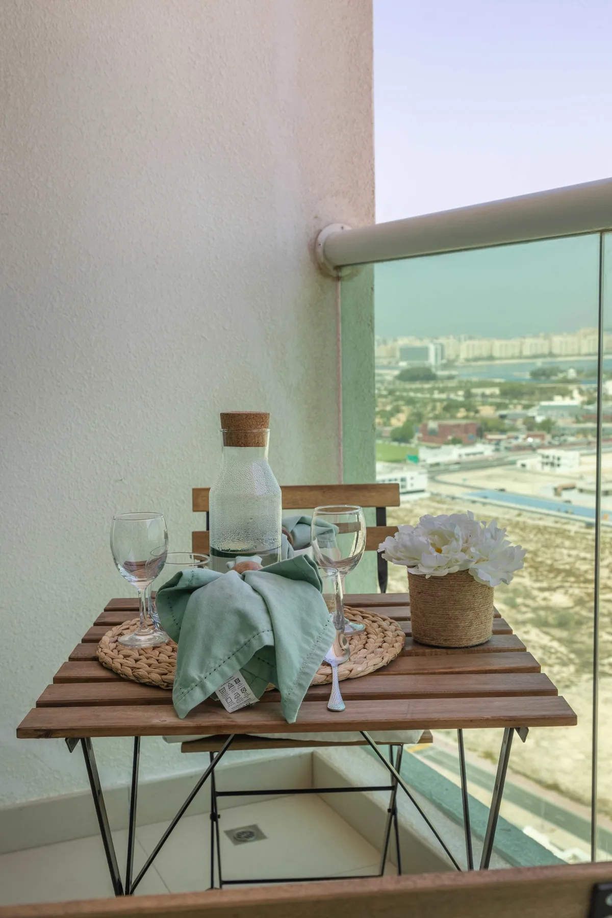 Una mesa de balcón está preparada con bebidas y flores junto a una barandilla de vidrio con vistas a la ciudad.