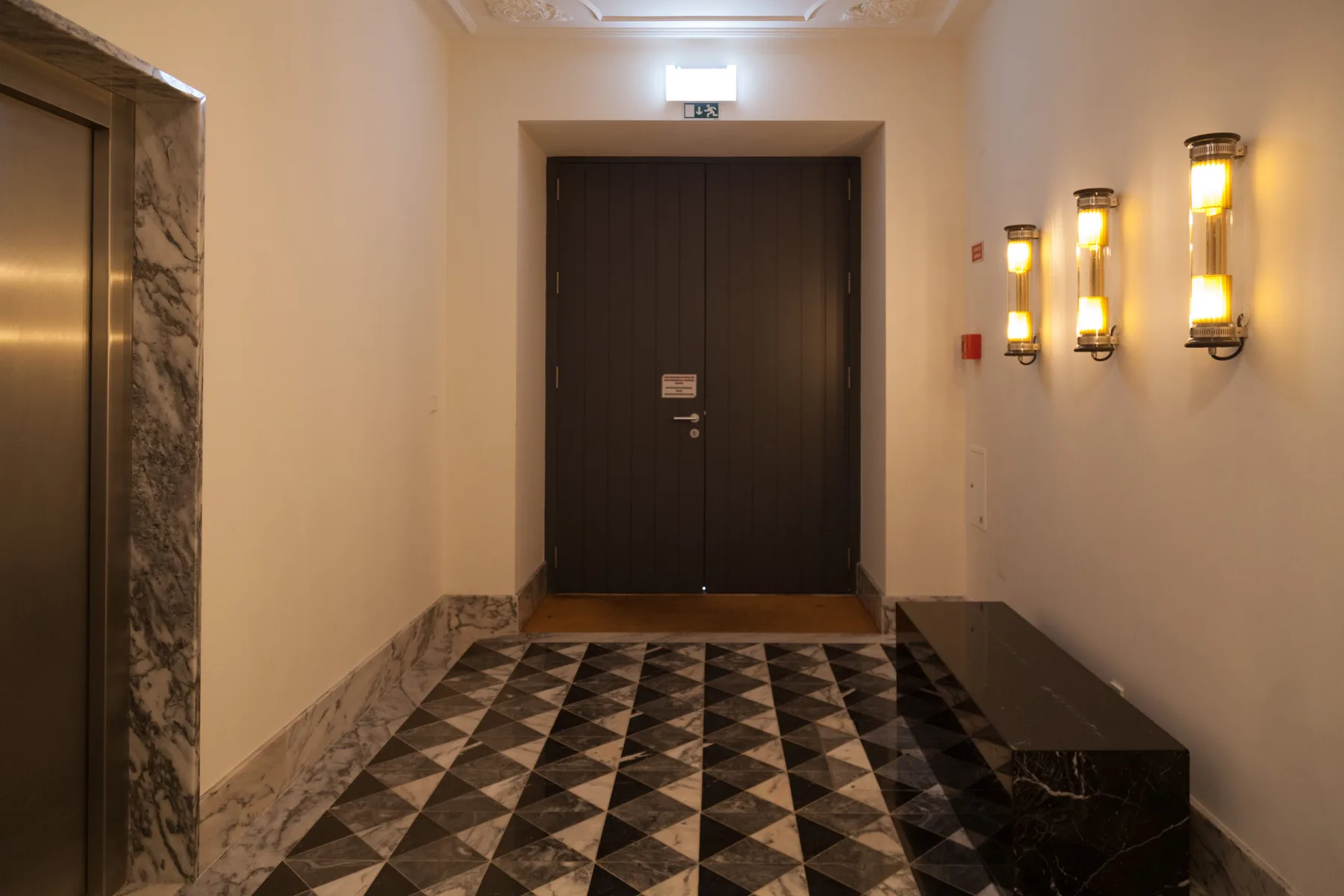 An interior hallway with a dark door, patterned floor, and wall lights.