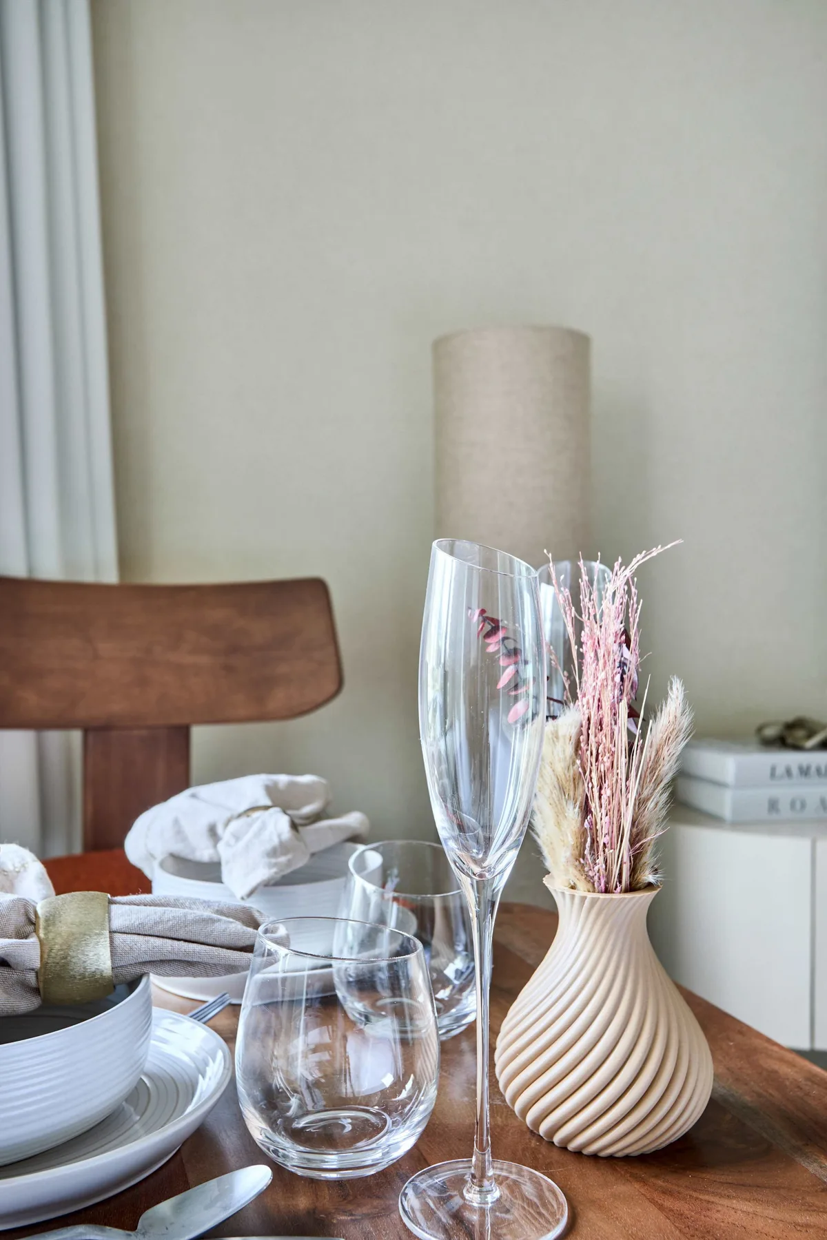 Una mesa de comedor está puesta con cristalería, ropa de mesa doblada y un jarrón con flores secas.