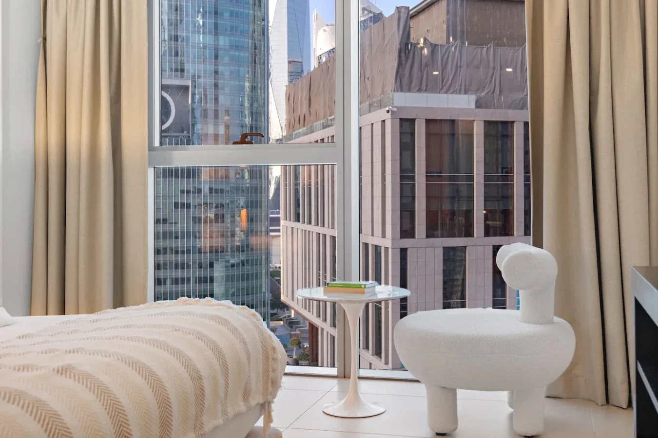 Un dormitorio con una cama junto a una gran ventana con vistas a edificios de la ciudad.