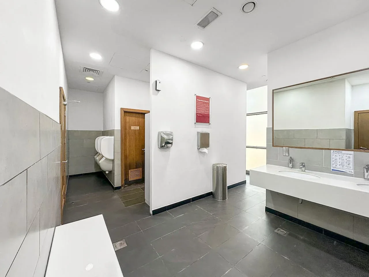 Un baño público luminoso con paredes de azulejos, una encimera con lavabos y una zona de inodoro.