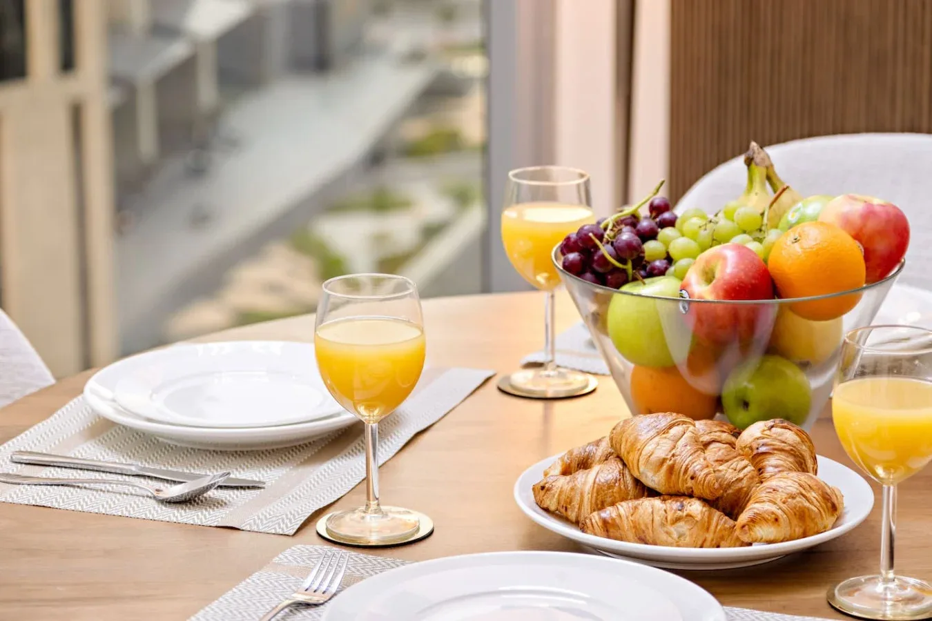 Una mesa de comedor está preparada con platos, zumo, fruta y cruasanes.