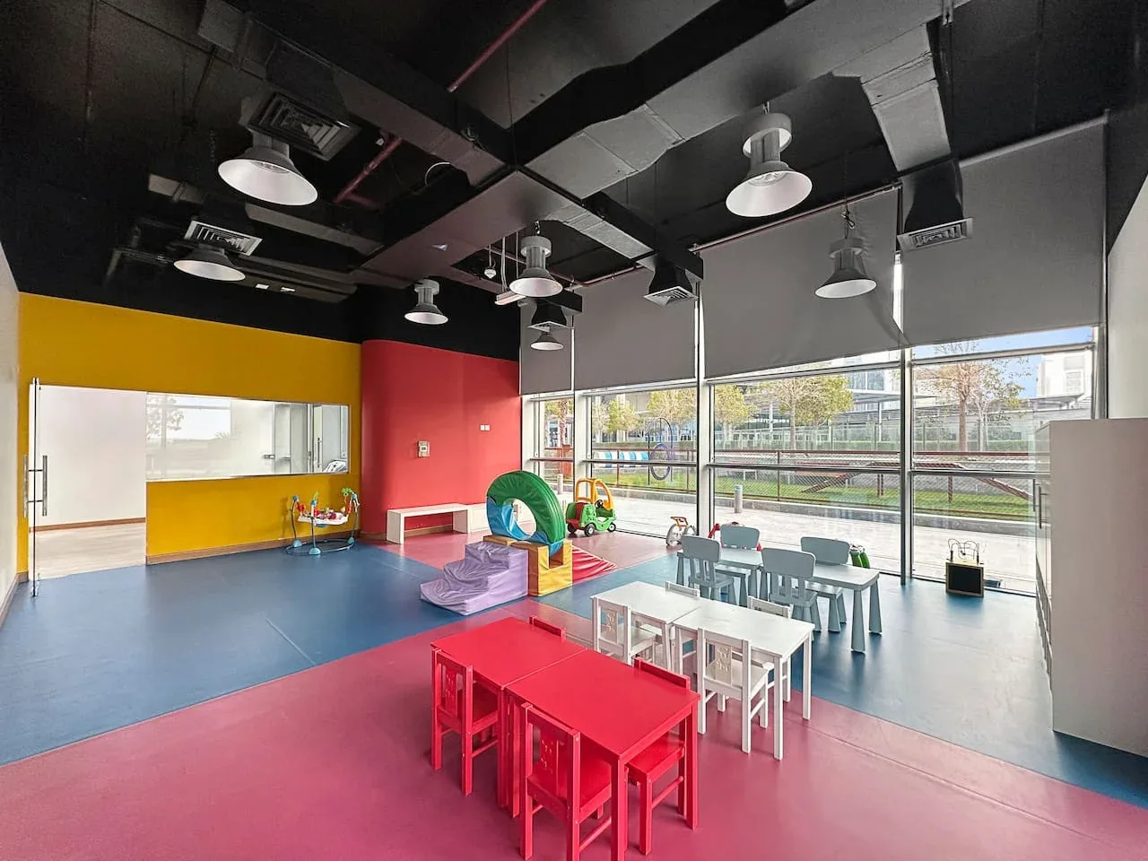 Una sala de juegos colorida con mesas infantiles y juegos junto a grandes ventanales.
