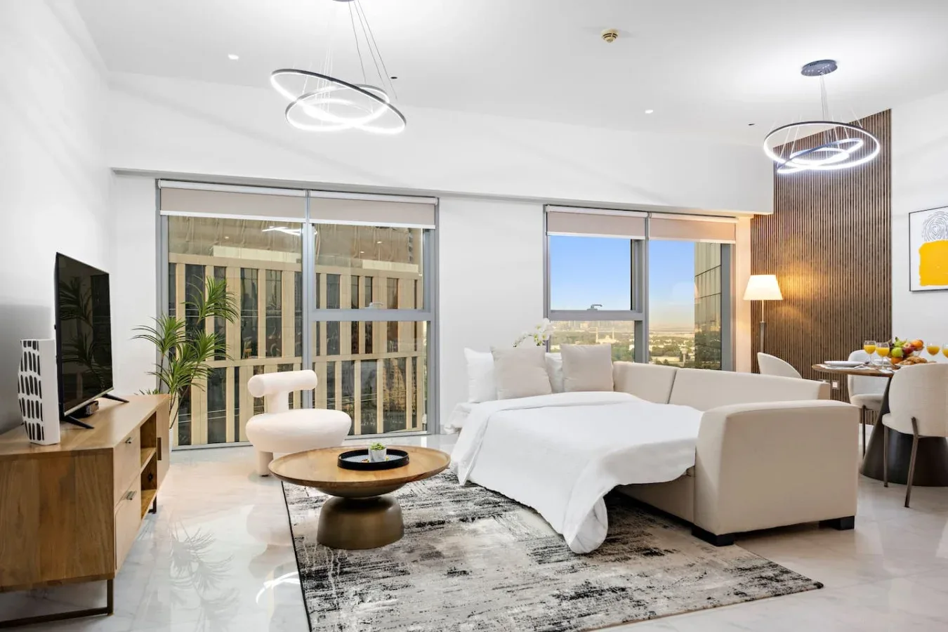 Un dormitorio luminoso con una cama grande, asientos y ventanas con vistas a edificios cercanos.