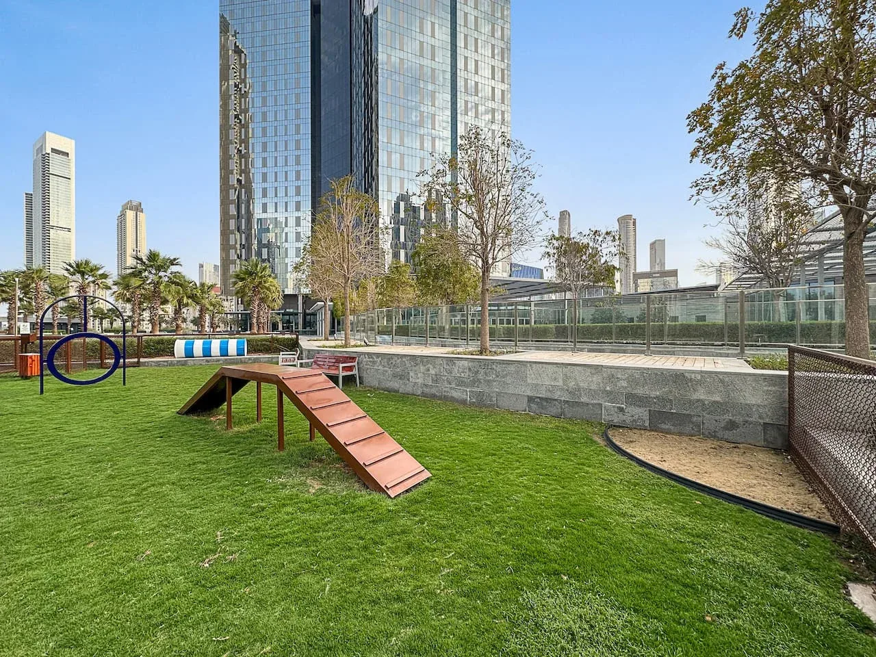 Una zona de juegos al aire libre con césped, una rampa de madera y edificios altos de cristal al fondo.