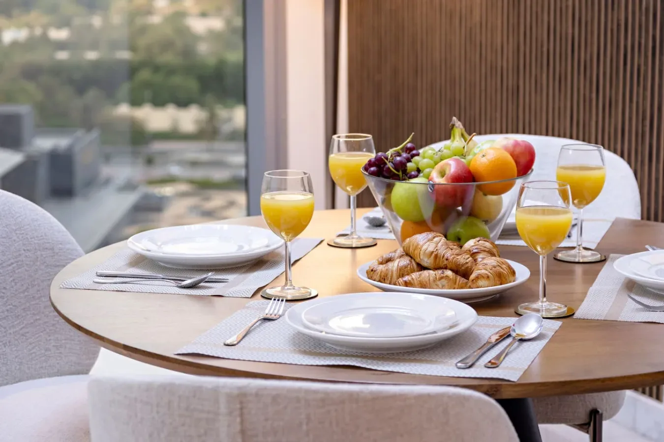 Una mesa de comedor está preparada con fruta, bollería, platos y vasos de zumo junto a una ventana.