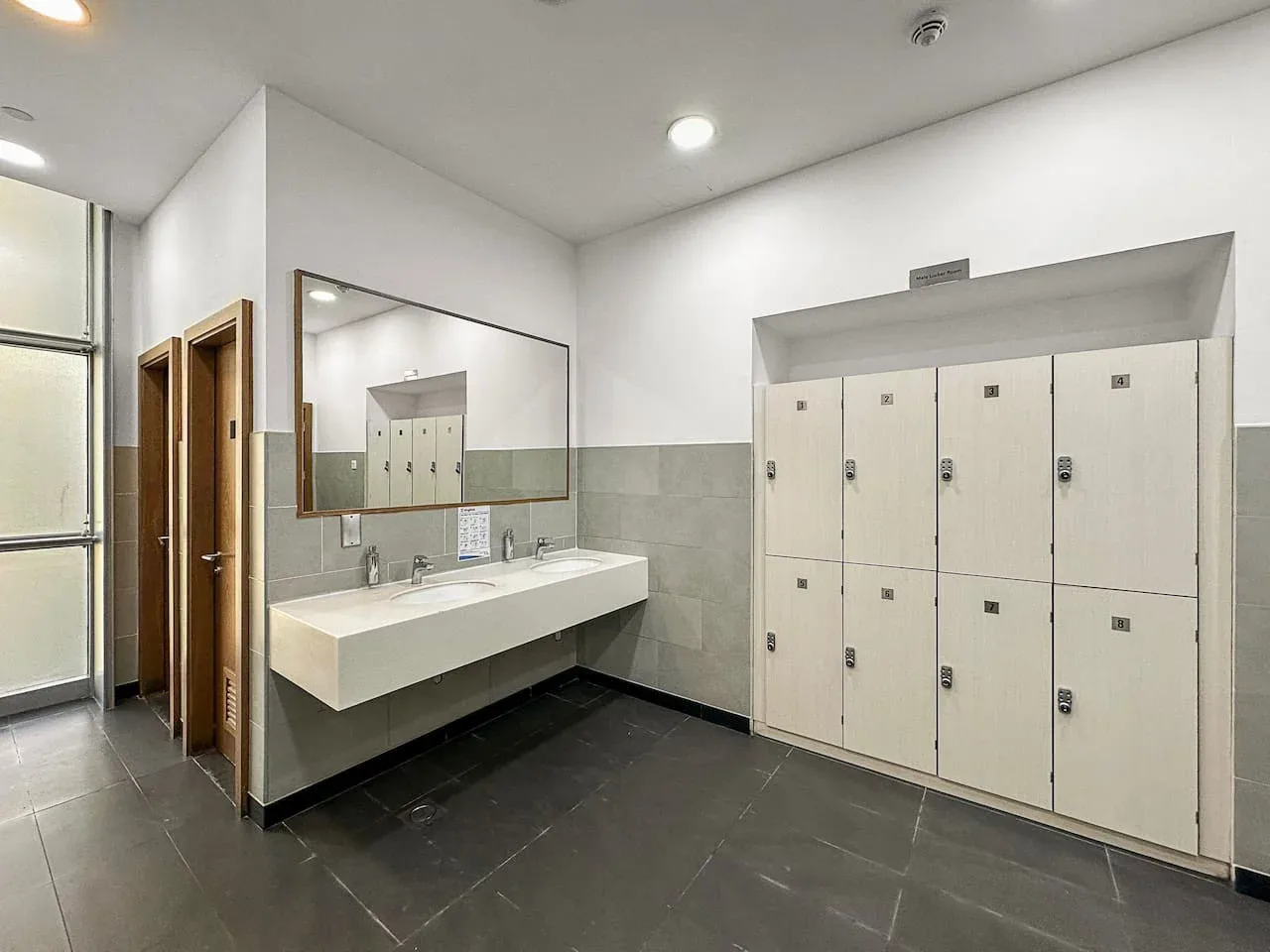 Un baño luminoso con una encimera larga con lavabos, un espejo y taquillas.