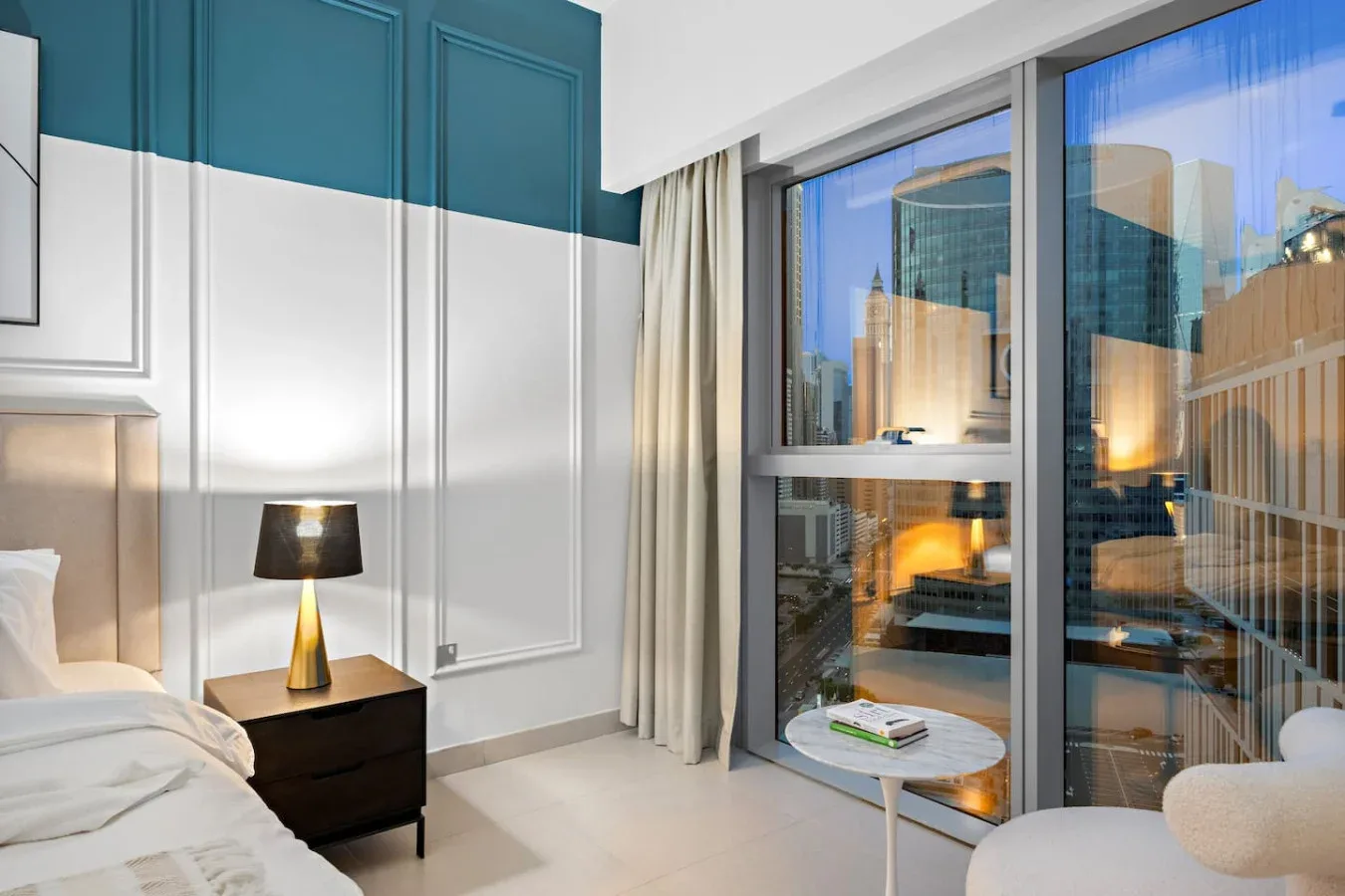 Un dormitorio con cama, mesita de noche y grandes ventanas con vistas a edificios cercanos.