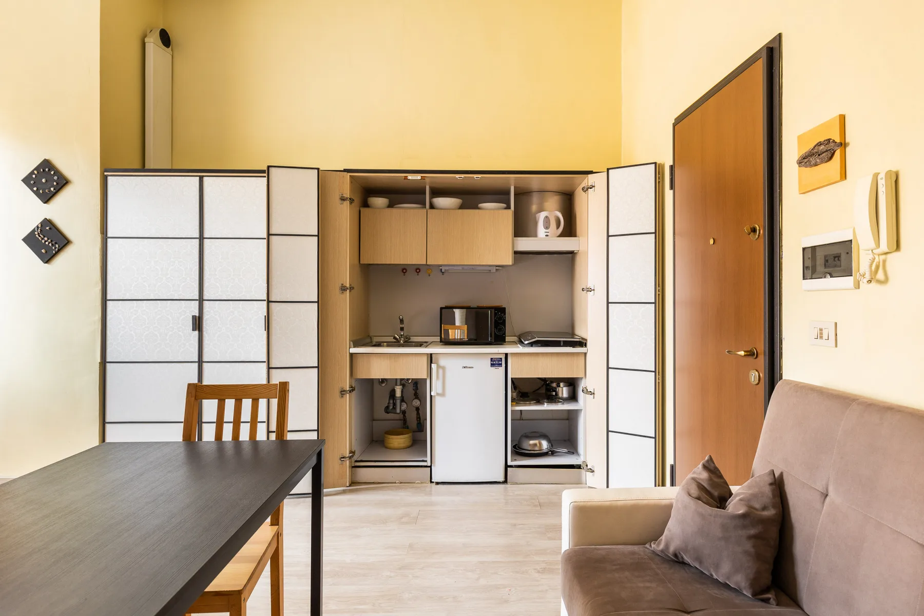 Una pequeña cocina se encuentra entre una mesa de comedor y un sofá en una habitación luminosa.